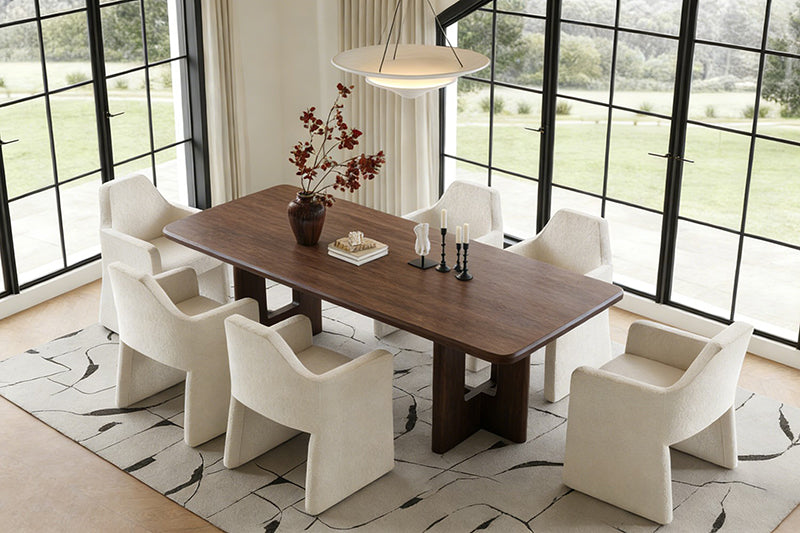A rendering of a modern minimalist dining room: a walnut dining table paired with cream-colored upholstered chairs, set against large floor-to-ceiling windows overlooking an outdoor lawn.