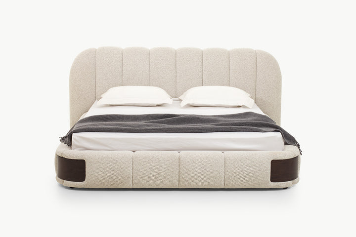 Luxurious upholstered bed with tall scalloped channel-tufted headboard in soft beige textured fabric and padded segmented base with dark wood side accents