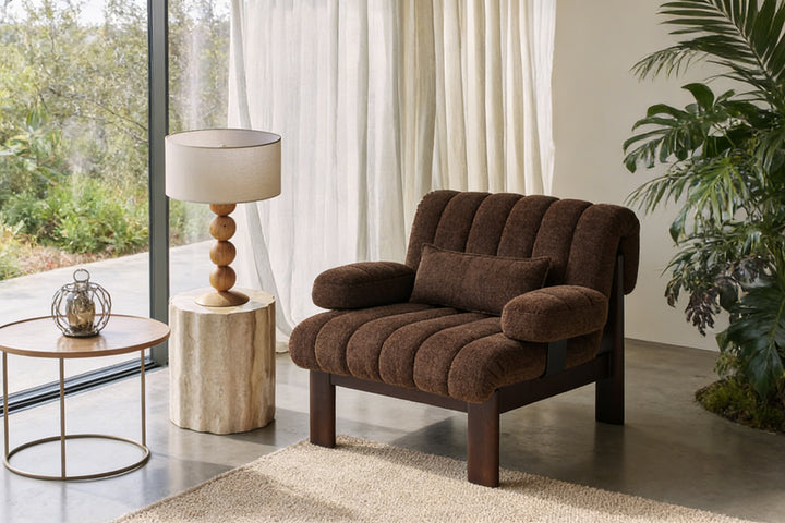 Warm chocolate boucle Coco Armchair beside lamp, plush enveloping shape, ideal cozy spot for coffee and books